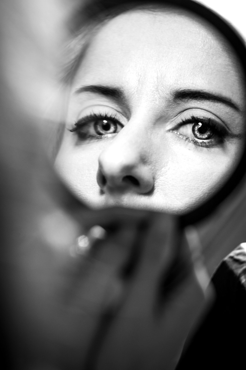 A greyscale image looking over the shoulder of a young woman into a mirror, where her pale eyes stare back at the viewer intensely