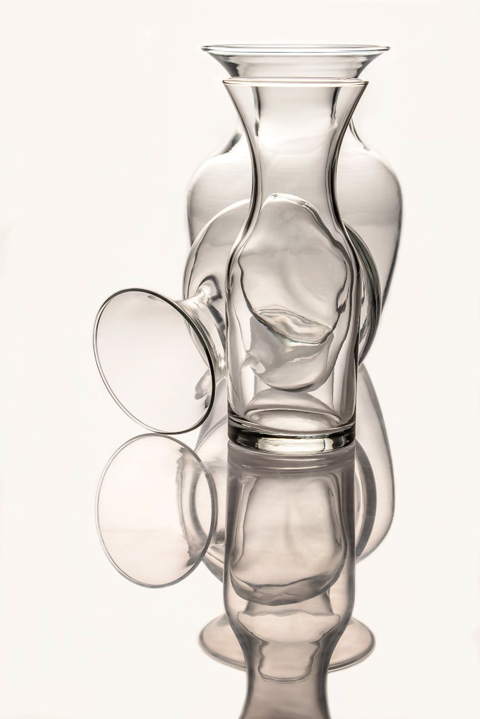 Three glass decanters decreasing in size, arranged in a single line, with the middle one tipped to open with its mouth on the left-side of the table--white background
