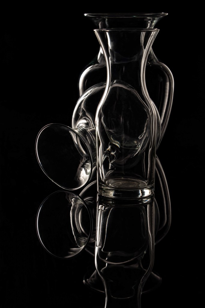 Three glass decanters decreasing in size, arranged in a single line, with the middle one tipped to open with its mouth on the left-side of the table--black background
