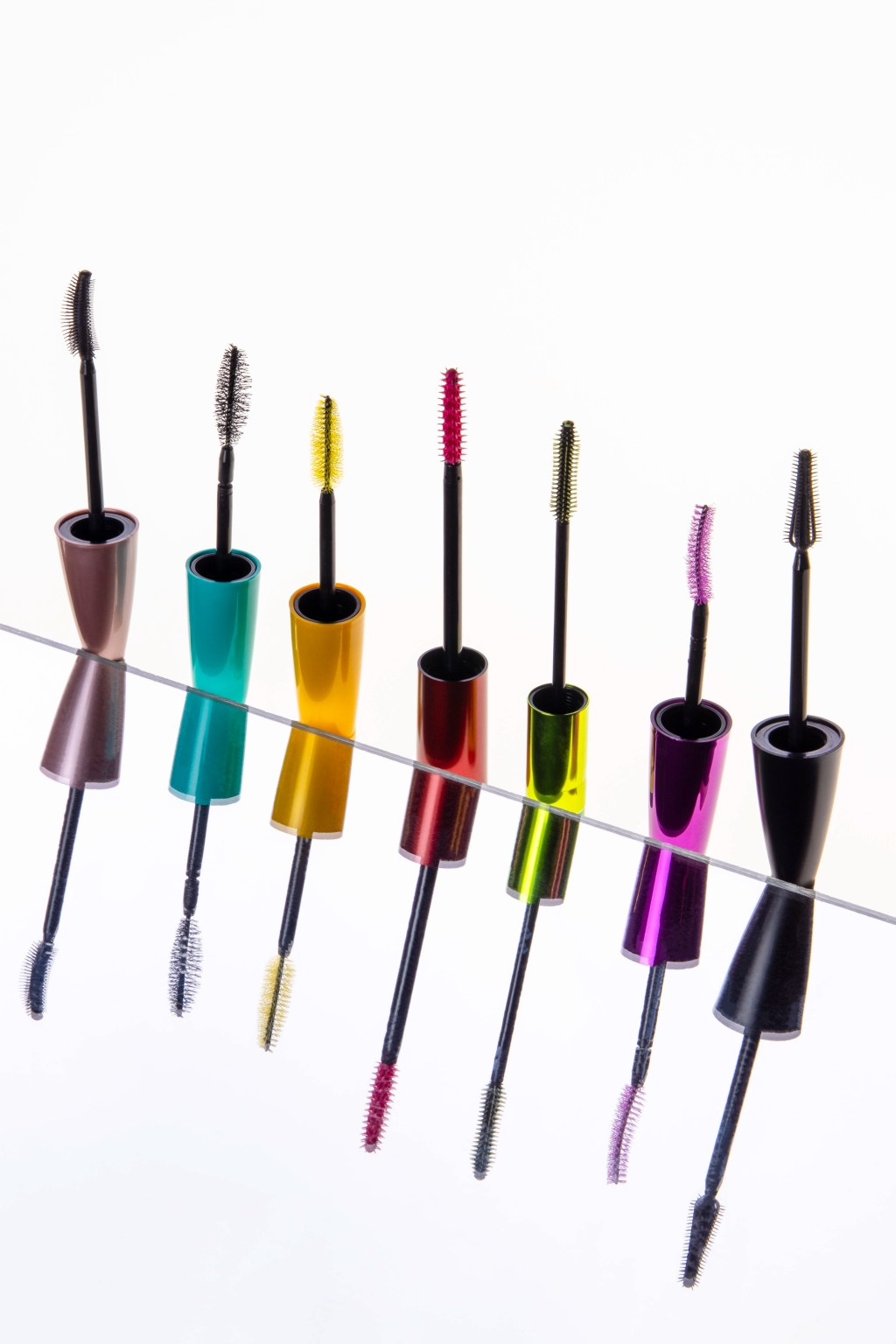 7 mascara wands line up across a white background in the colors lavender, aqua, yellow, red, lime, fuschia, and black. They are reflected in a mirror below.