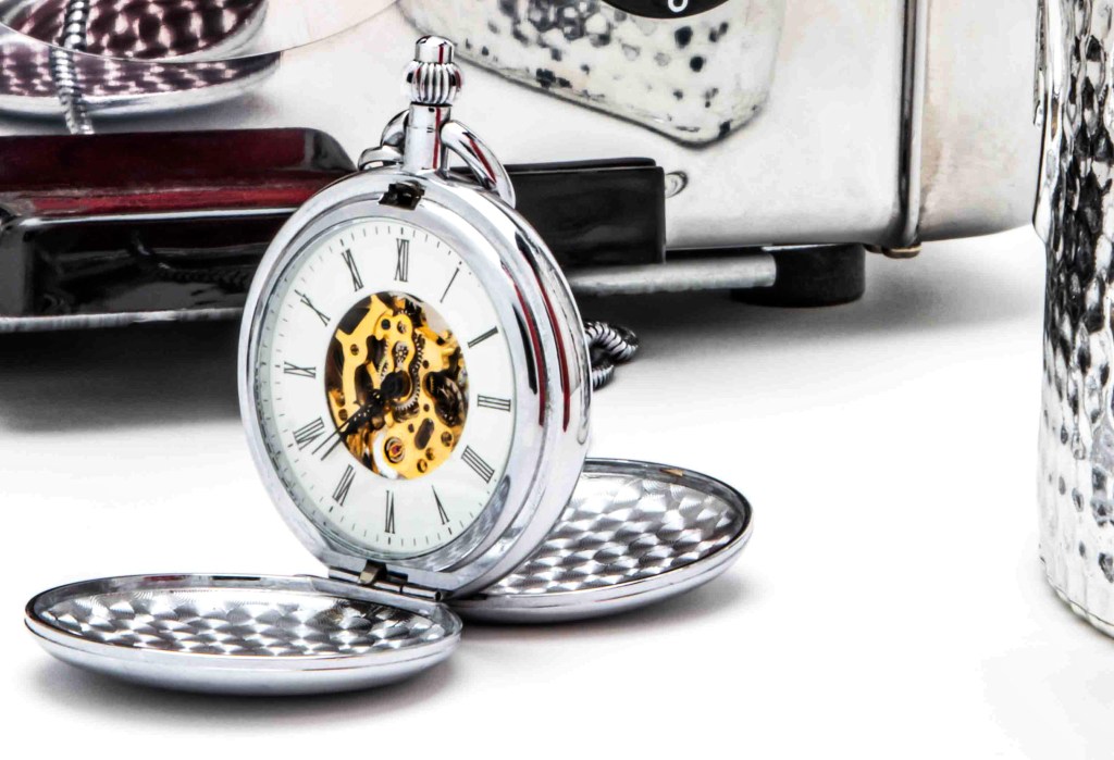 A silver pocket watch lies open on a white table, reflected in a metal surface behind and another to the right