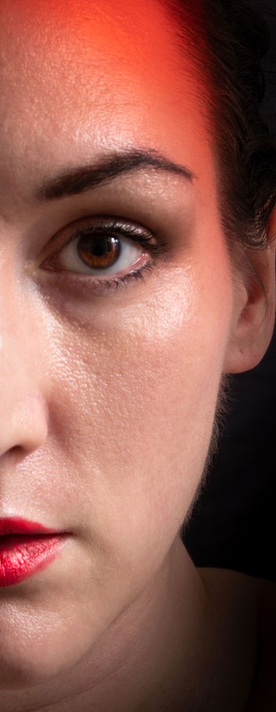 Left half of a woman's face, dark hair, dark eye, red lips