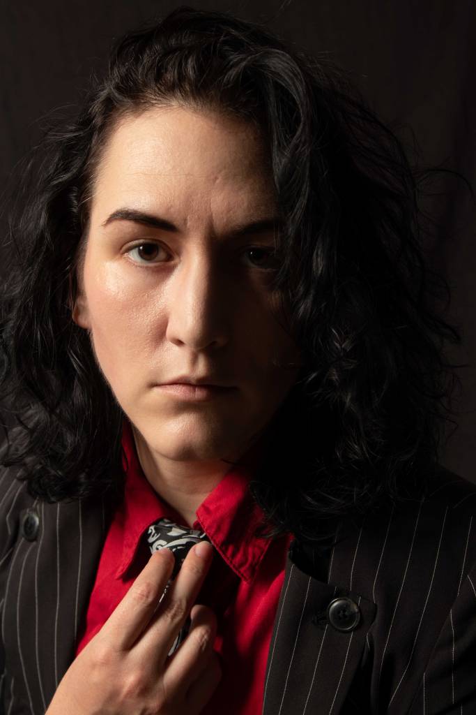 fair skin androgynous person in a red shirt, silver and black skull tie and pinstripe suit jacket