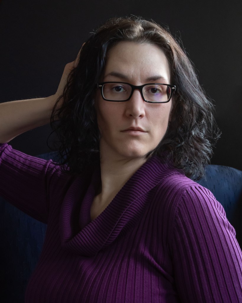 Self portrait, fair woman with dark shoulder length hair and dark eyes wearing a purple shirt and glasses