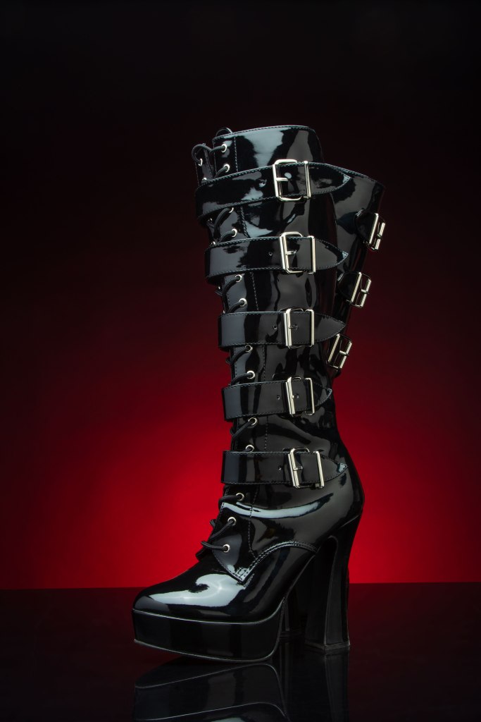 Vinyl knee high black fetish boots with 5 buckles backlit with a red light on a black background